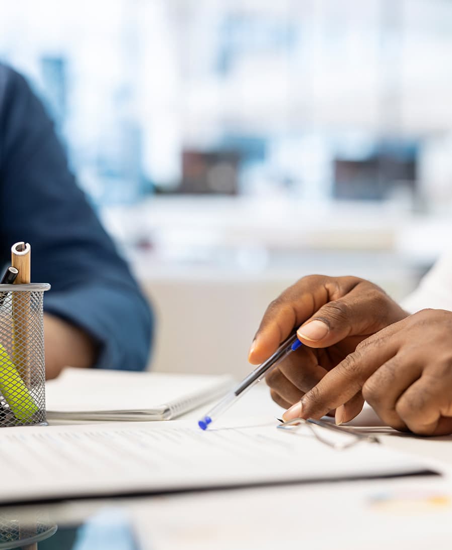 A hand holding a pen over paperwork on a desk, beside a second person